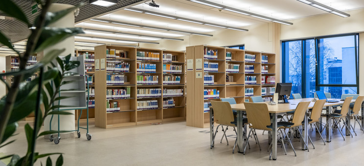 Na zdjęciu wnętrze Centrum Informacyjno-Bibliotecznego. Po prawej stronie przy oknach rząd połączonych stolików otoczony krzesłami. W tle regały z książkami o kolorowych okładkach. Wszystkie meble mają odcień jasnego drewna.